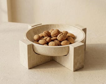 May include: A beige stone bowl with a smooth, circular basin supported by four rectangular, block-like legs. The bowl is filled with a collection of brown, textured walnuts. The entire object is crafted from a consistent, light-colored natural stone material.
