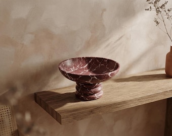 May include: A deep burgundy marble bowl with prominent white veining, featuring a wide, shallow basin supported by a thick, tiered, circular pedestal base. The stone surface displays a polished finish with natural patterns throughout the material.