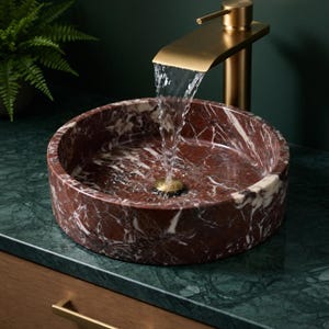 May include: A round, deep red marble vessel sink with white veining, featuring a brushed gold waterfall faucet. Water flows from the faucet into the center of the basin, which has a matching gold drain. The sink sits on a dark green marble countertop.