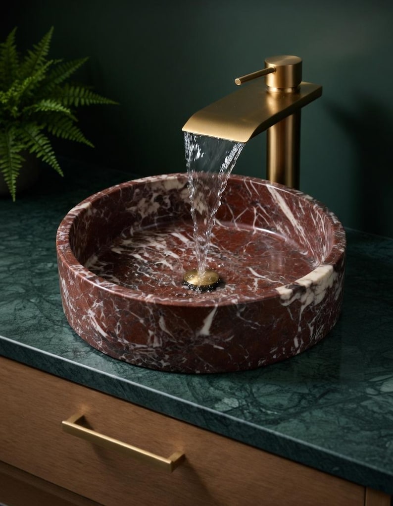 May include: A round, deep red marble vessel sink with white veining, featuring a brushed gold waterfall faucet. Water flows from the faucet into the center of the basin, which has a matching gold drain. The sink sits on a dark green marble countertop.