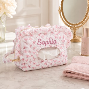 May include: A white quilted tissue box cover featuring a pink bow pattern and a ruffled top edge. The name Sophia is embroidered in pink script on the front. The rectangular fabric case has a central opening for tissues and a side handle.