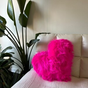 May include: A bright pink heart-shaped pillow made of fluffy faux fur. The pillow is sitting on a white couch.