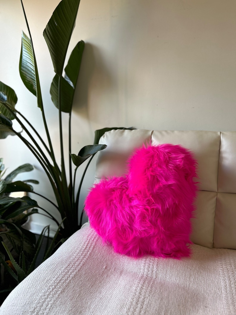 May include: A bright pink heart-shaped pillow made of fluffy faux fur. The pillow is sitting on a white couch.