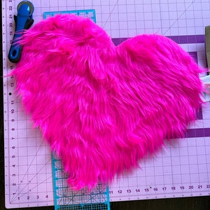 May include: A pink heart-shaped piece of faux fur fabric on a cutting mat with a ruler and rotary cutter.
