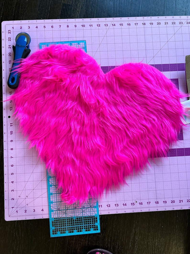 May include: A pink heart-shaped piece of faux fur fabric on a cutting mat with a ruler and rotary cutter.