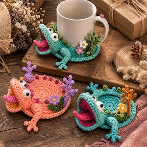 May include: Three crocheted coasters shaped like axolotls, one in peach and two in teal. Each features a wide-open mouth with a pink tongue, white eyes, and small floral or coral-like accents. One teal coaster holds a white ceramic mug.
