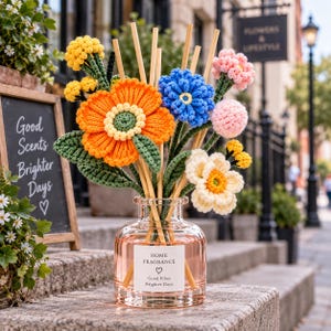 May include: A clear glass home fragrance diffuser bottle filled with pink liquid and wooden reeds, topped with a bouquet of crocheted flowers in orange, blue, yellow, pink, and white. The label reads: HOME FRAGRANCE, Good Vibes, Brighter Days.