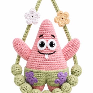 Puede incluir: Un personaje de estrella de mar rosa con grandes ojos negros y una pequeña lengua blanca, que lleva pantalones cortos de color verde claro con patrones de flores moradas. La figura cuelga de correas de ganchillo verde claro con flores.
