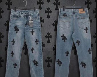 May include: Light blue denim jeans featuring a repeating pattern of black cross-shaped patches. The jeans are displayed from both the front and back views, showing a button fly, belt loops, and a tan leather-like brand patch on the rear.