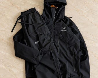 May include: A black Arc'teryx hooded jacket and matching cargo pants set. The jacket features a front zipper, a hood, and the white Arc'teryx logo on the chest. The pants include a drawstring waist, side cargo pockets, and the white Arc'teryx logo on the leg.