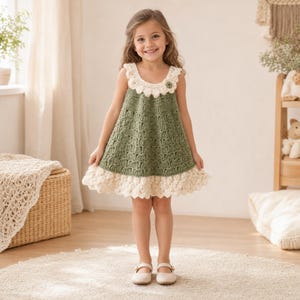 May include: A sleeveless crochet dress featuring an olive green bodice with a textured shell stitch pattern, accented by a cream-colored scalloped collar and a matching wide ruffled hem. The dress includes a small decorative flower on the neckline.
