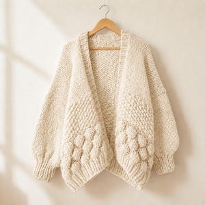 Può includere: Un cardigan in maglia grossa color bianco sporco con apertura frontale, maniche lunghe con polsini a coste e un motivo testurizzato di punti a bolle in rilievo sui pannelli anteriori inferiori. Il capo è appeso a una gruccia di legno.