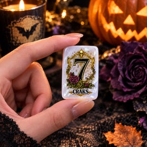 May include: A rectangular white mother-of-pearl token featuring a gold ornate frame with a skull at the top, a large black number 7 in the center, pink and green roses at the base, and the word CRAKS printed in black lettering at the bottom.