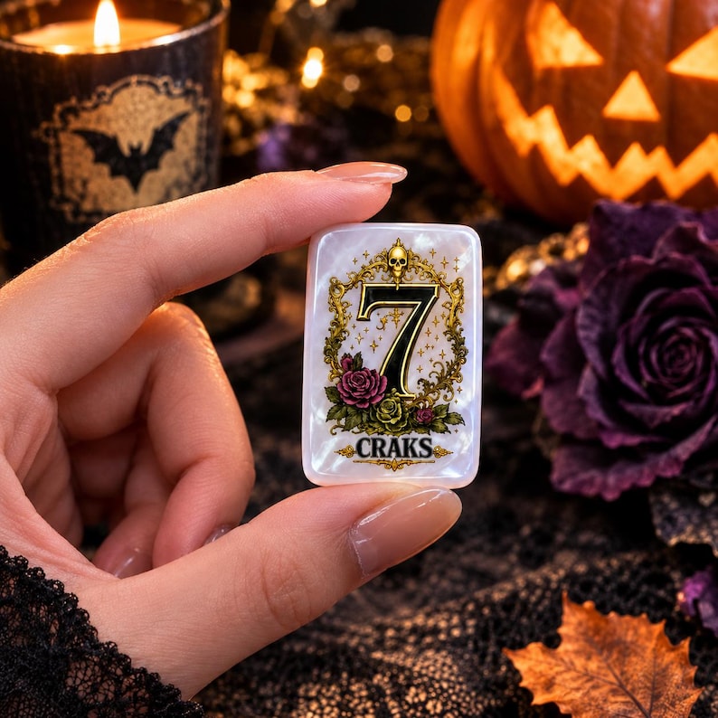 May include: A rectangular white mother-of-pearl token featuring a gold ornate frame with a skull at the top, a large black number 7 in the center, pink and green roses at the base, and the word CRAKS printed in black lettering at the bottom.