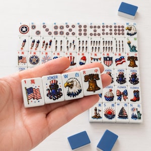 May include: A set of white mahjong tiles featuring patriotic American-themed illustrations including flags, eagles, fireworks, and the Liberty Bell. Four tiles are held in a row, displaying a flag, a joker octopus, an eagle, and a bell.