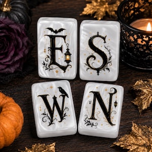 May include: Four rectangular white tiles with a pearlescent finish, each featuring a large black letter forming the word E S W N. The letters are decorated with gold stars, black filigree, a bat, a lantern, a crow, and a ghost illustration.