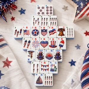 May include: A collection of white rectangular mahjong tiles arranged in a pyramid shape, featuring colorful patriotic American-themed illustrations including fireworks, flags, a liberty bell, a top hat, a sailboat, and the Statue of Liberty.