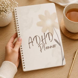 Op de afbeelding: Een spiraalgebonden planner met een crèmekleurige kaft voorzien van een zachte, beige bloemenillustratie in aquarel-stijl en de donkerbruine tekst "ADHD Planner" in het midden gedrukt. Het notitieboek heeft een gouden metalen spiraal aan de linkerkant.