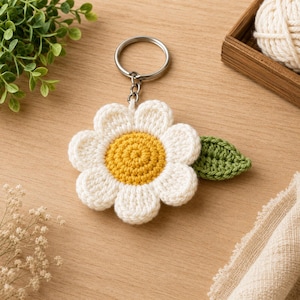 Könnte beinhalten: Ein gehäkelter Gänseblümchen-Schlüsselanhänger mit einer gelben Mitte, weißen Blütenblättern und einem einzelnen grünen Blatt an der Seite. Die Blume ist an einem silberfarbenen Metallring befestigt, der als Halterung für Schlüssel dient.