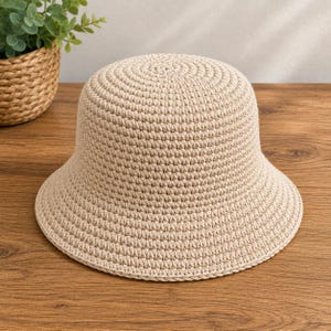Può includere: Un cappello a secchiello beige caratterizzato da un motivo a uncinetto denso e uniforme. Il cappello presenta una corona arrotondata e una tesa inclinata verso il basso, realizzato in un filato continuo di colore neutro con una trama costante.