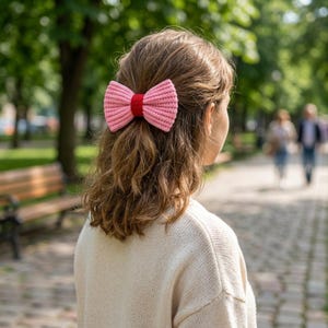Könnte beinhalten: Ein Haaraccessoire bestehend aus einer großen, gerippten, hellrosa Schleife mit einem einfarbigen roten Mittelband. Die Schleife ist aus einem strukturierten Strickmaterial gefertigt und im Haar befestigt, was einen auffälligen, farbigen Akzent setzt.