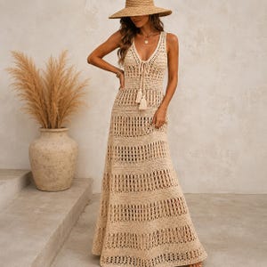 Könnte beinhalten: Ein ärmelloses, beiges Häkel-Maxikleid mit tiefem V-Ausschnitt, Kordelzug und Quasten. Das Kleid ist aus abwechselnden horizontalen Bändern mit durchbrochenen und festen Häkelmaschen gefertigt und reicht bis zum Boden.