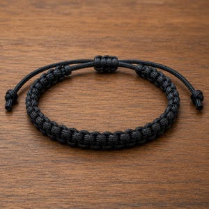 May include: A black paracord bracelet featuring a woven knot pattern. The bracelet includes an adjustable sliding knot closure mechanism with two thin cord ends, each finished with a small knotted stopper at the very tip of the cord.