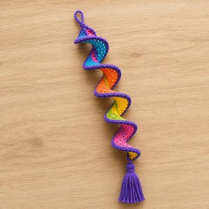 May include: A spiral-shaped crochet ornament featuring a vibrant rainbow color gradient of purple, blue, orange, yellow, green, and pink yarn. The piece includes a small loop at the top for hanging and a purple tassel attached to the bottom end.