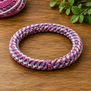 Könnte beinhalten: Ein kreisförmiges Armband aus geflochtener Kordel in einer bunten Farbpalette aus Magenta, Lavendel, Violett und Weiß. Das Accessoire zeichnet sich durch eine durchgehende, fest gewebte Textur mit gleichmäßiger Dicke über das gesamte Band aus.