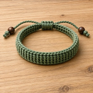 Könnte beinhalten: Ein salbeigrünes Armband aus gewebter Kordel mit einem dicken, strukturierten Makramee-Muster. Das Armband verfügt über einen verstellbaren Schiebeknotenverschluss und zwei dunkelbraune Holzperlen an den Enden der Zugkordeln für ein schlichtes Design.