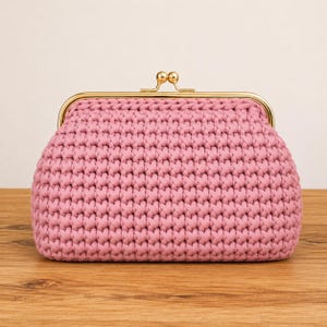 Könnte beinhalten: Eine altrosa gehäkelte Clutch mit einem dicken, strukturierten Strickmuster. Die Tasche wird mit einem goldfarbenen Metall-Schnappverschluss gesichert, der zwei kleine, runde, polierte goldene Kugeln an der Oberseite zum Öffnen und Schließen besitzt.