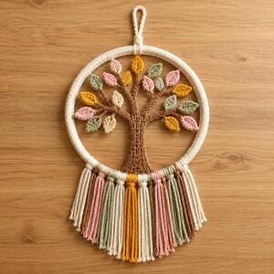 Könnte beinhalten: Ein kreisförmiger Wandbehang mit einem braunen gehäkelten Baum mit bunten Blättern in Rosa, Gelb, Grün und Creme. Der Baum ist in einen weißen gehäkelten Ring mit einer Aufhängeschlaufe und einer Fransenborte aus Garn am unteren Rand eingefasst.