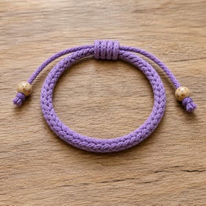 Könnte beinhalten: Ein violettes Armband aus geflochtener Kordel mit einem Schiebeknoten-Verschluss. Die beiden Enden der Kordel sind mit kleinen, runden, beigefarbenen Perlen versehen, die dunkelbraune Sprenkel aufweisen und einen dekorativen Akzent setzen.