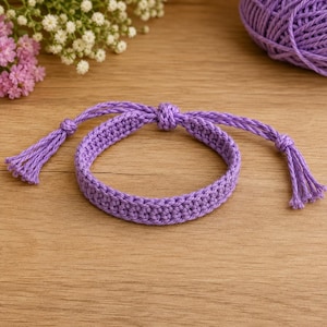 Könnte beinhalten: Ein lavendelfarbenes, gewebtes Armband mit einem zentralen geflochtenen Band, verstellbaren Schiebeknoten und zwei herabhängenden Quastenenden. Das Armband besteht aus einer durchgehenden Kordel und weist ein gleichmäßiges, strukturiertes Maschenbild auf.