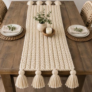 May include: A rectangular cream-coloured crocheted table runner featuring a thick ribbed texture and four large tassels attached to each of the two short ends. The runner is made of chunky yarn and displays a consistent, uniform knit pattern.
