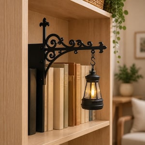 May include: A black metal wall-mounted bracket with ornate scrollwork and a fleur-de-lis top finial. A small black lantern with a clear glass pane and a warm glowing light bulb hangs from the bracket by an S-hook, positioned next to a row of books.