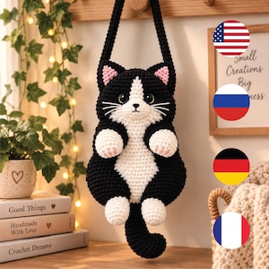 Könnte beinhalten: Eine schwarz-weiße, katzenförmige Häkeltasche mit langem Trageriemen. Die Katze hat eine weiße Brust, ein weißes Gesicht und weiße Pfoten, rosa Innenohren, schwarze Schnurrhaare und große dunkle Augen. Die Tasche hat eine strukturierte Garnoberfläche.