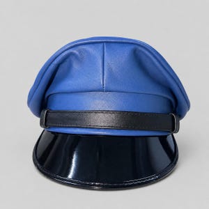 May include: A bright blue cap featuring a smooth, matte finish on the crown. A black leather-like strap is positioned horizontally across the front, secured by a buckle. The cap includes a wide, curved, glossy black visor at the base.