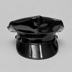 May include: A black patent leather-look military-style cap with a high-gloss finish. The hat features a structured crown, a horizontal band across the front, and a curved visor. The material has a reflective surface with visible stitching details.