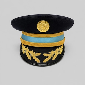 May include: A dark blue military service cap featuring a light blue band, a gold-coloured chin strap, and a gold-coloured emblem on the front. The black visor is decorated with gold-coloured embroidered oak leaf patterns on both sides.