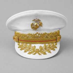 Könnte beinhalten: Eine weiße Militärmütze mit Schirm, die vorne mittig mit einem goldfarbenen Emblem aus Adler, Weltkugel und Anker versehen ist. Die Mütze verfügt über einen goldenen Kinnriemen mit rotem Streifen und goldene Blattstickereien am Schirm.