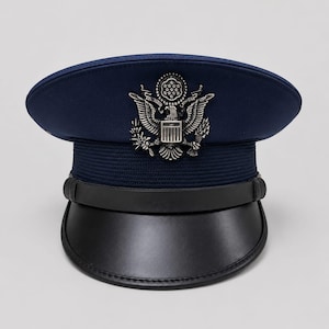 May include: A dark blue service cap featuring a silver-toned metal emblem of an eagle with outstretched wings, a shield, and a banner. The cap includes a textured blue band, a black leather chin strap, and a black leather visor brim.