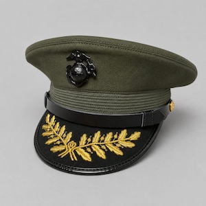 May include: An olive green military service cap featuring a black leather chin strap and a black visor adorned with gold embroidered oak leaves. A black metal Eagle, Globe, and Anchor emblem is centered on the front of the olive green crown.