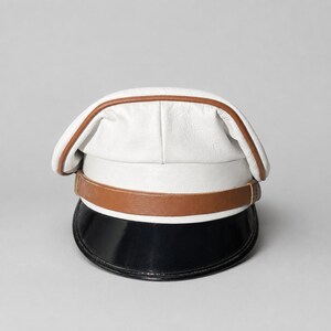 May include: A white leather captain hat featuring a brown leather band across the front and a glossy black visor. The crown of the hat is constructed from white leather panels with brown leather piping along the top edge and a structured band.