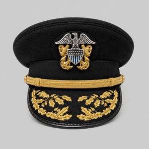 May include: A black military-style service cap featuring a prominent silver and gold eagle emblem centered above a gold chin strap. The visor is decorated with gold embroidered oak leaf clusters, and the cap has a structured, rounded crown.