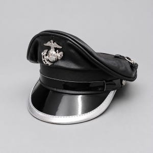 Könnte beinhalten: Eine schwarze Militärmütze aus Leder mit einem silberfarbenen Emblem aus Adler, Weltkugel und Anker auf der Vorderseite. Die Mütze hat einen glänzenden Schirm aus schwarzem Lackleder mit silbernem Rand und einen passenden Kinnriemen.