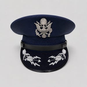 May include: A dark blue military service cap featuring a silver-toned eagle emblem centred on the front. The cap includes a black chin strap and a visor decorated with silver embroidered cloud and lightning bolt motifs on both sides.