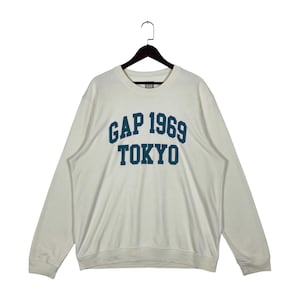 Peut inclure: Un sweat-shirt à manches longues blanc cassé avec une encolure ronde et un cintre noir. L'avant présente le texte GAP 1969 TOKYO en lettres capitales bleu sarcelle foncé. Le vêtement est doté de poignets et d'un ourlet côtelés.