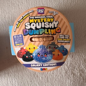 以下が含まれることがあります： Mystery Squishy Dumplingというおもちゃの丸い紙製パッケージ、ギャラクシーエディション。タンカラーのラベルには、様々な餃子のカラフルなイラスト、QRコード、「The Original Viral Mystery Squishy Dumpling」というテキストが記載されています。