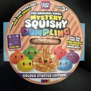 Könnte beinhalten: Eine runde Kartonverpackung für ein Mystery Squishy Dumpling Spielzeug. Das Etikett zeigt den Text "The Original Viral Mystery Squishy Dumpling" und "Golden Stuffed Edition!" mit Abbildungen von bunten, lächelnden Knödel-Figuren.
